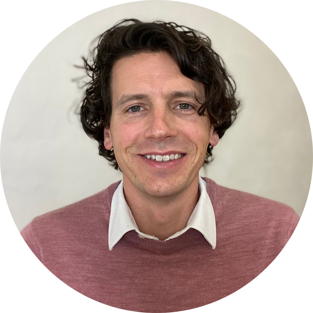 A circular headshot of a smiling man with wavy dark hair, wearing a mauve sweater over a white collared shirt against a neutral white background.